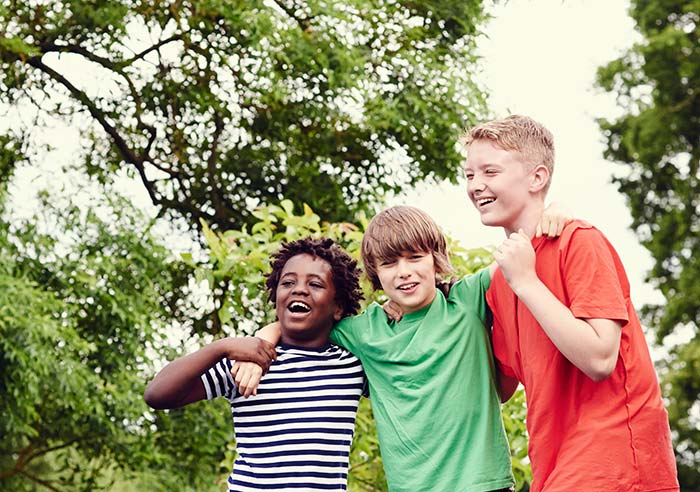 Three young boys hugging each other. They are outside and surrounded by trees.